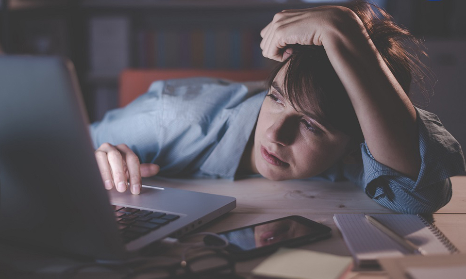 Persona stanca che lavora al computer di notte, con telefono e quaderno sul tavolo.