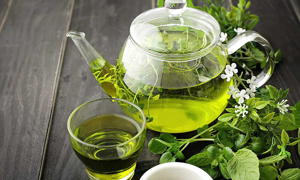 Teiera di vetro con tè verde, tazza piena, e foglie di erbe fresche su tavolo di legno nero.