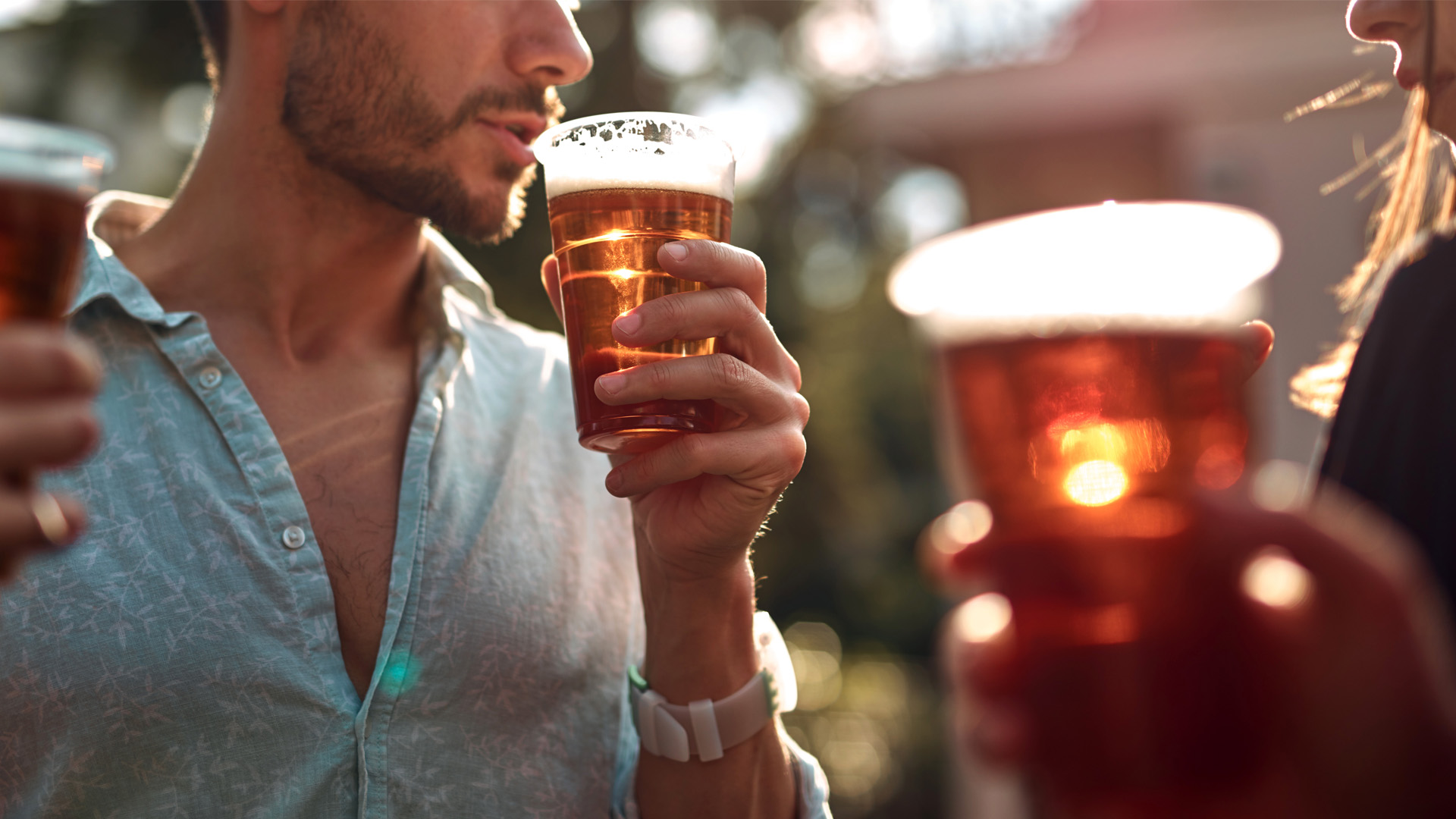 Persona che beve birra all'aperto, sole riflesso nel bicchiere di plastica in una scena estiva e conviviale.
